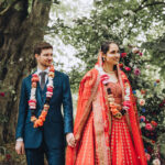 a groom and bride smile away from the camera lens looking out towards their guests. They're in a woodland setting, she's in a red lehenga and he's wearing a blue suit with flower garland
