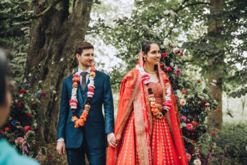 a groom and bride smile away from the camera lens looking out towards their guests. They're in a woodland setting, she's in a red lehenga and he's wearing a blue suit with flower garland