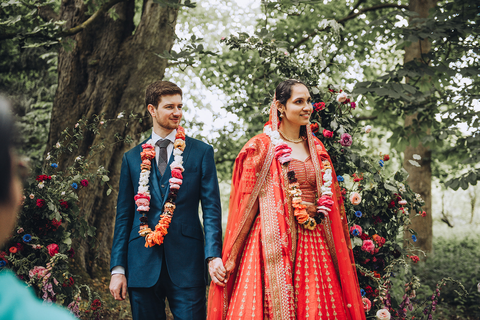 a groom and bride smile away from the camera lens looking out towards their guests. They're in a woodland setting, she's in a red lehenga and he's wearing a blue suit with flower garland