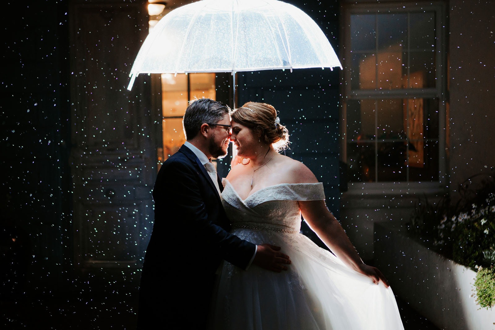 Beautiful low light photography of a groom and bride in the rain. She's holding a clear umbrella and the raindrops twinkle in the light. By Hannah Timm Photography in Cornwall