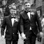 Two grooms walk hand in hand through petal confetti at an Essex wedding. By Ross Willsher Photography