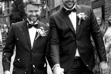 Two grooms walk hand in hand through petal confetti at an Essex wedding. By Ross Willsher Photography