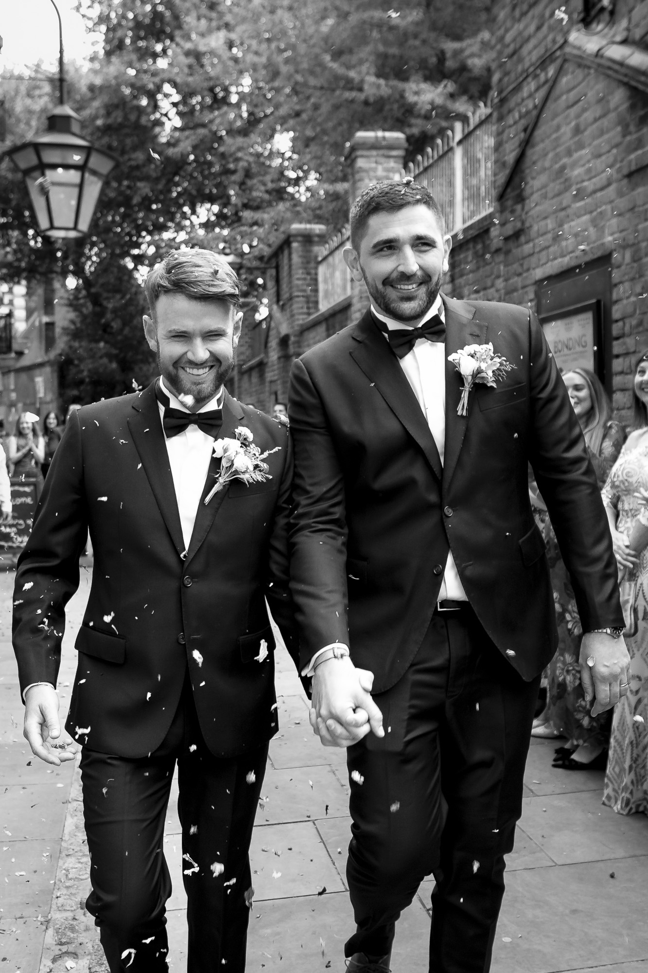 Two grooms walk hand in hand through petal confetti at an Essex wedding. By Ross Willsher Photography