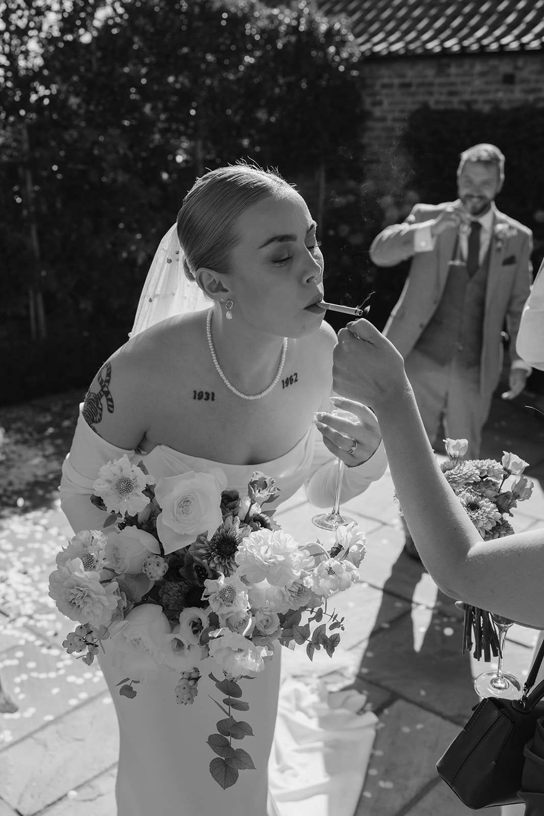 A tattooed bride has her cigarette lit for her by a guest. Black and white wedding photography by Joss Denham in Harrogate North Yorkshire 