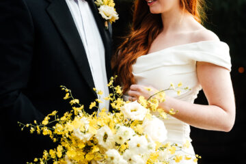 A model couple playing the role of groom and bride stand behind a bouquet of vibrant yellow spring flowers. He wears black tie and she's in an off-the-shoulder wedding dress