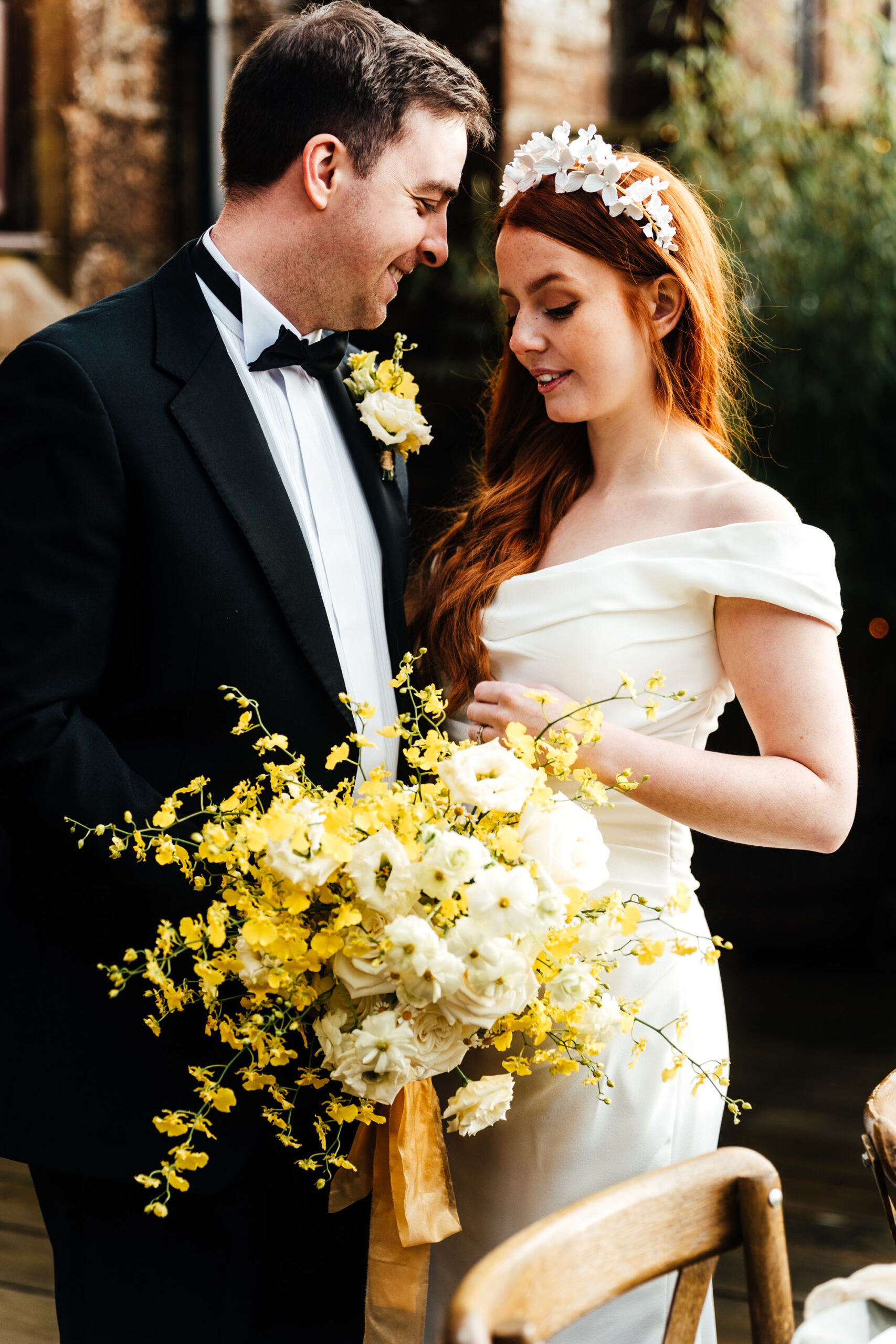 A model couple playing the role of groom and bride stand behind a bouquet of vibrant yellow spring flowers. He wears black tie and she's in an off-the-shoulder wedding dress