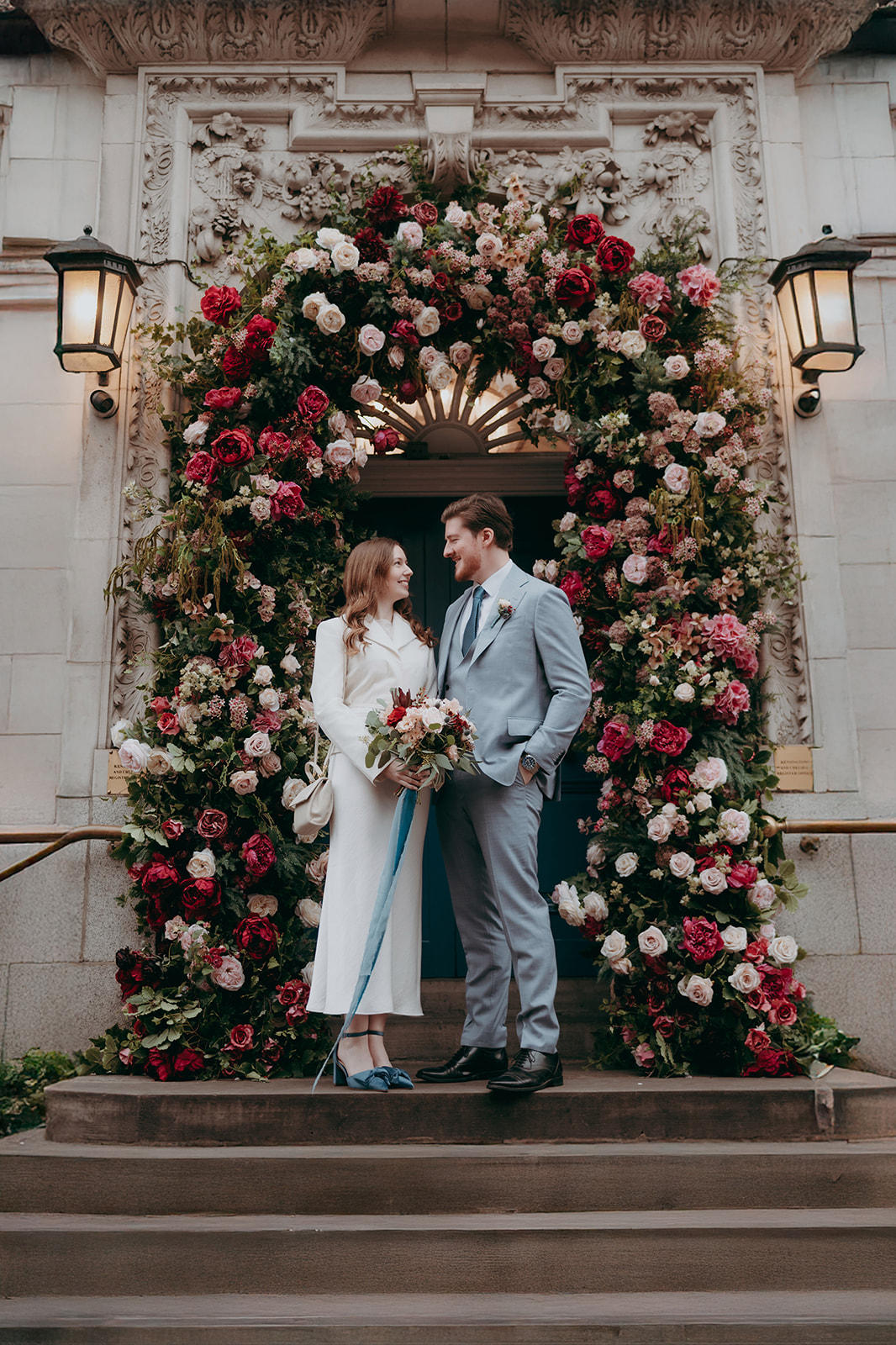 Portrait of newlyweds stood in front of a floral arch in reds, pinks and blush. She's wearing a slimline white dress and he's in a pale blue suit - by Damien Vickers Photography