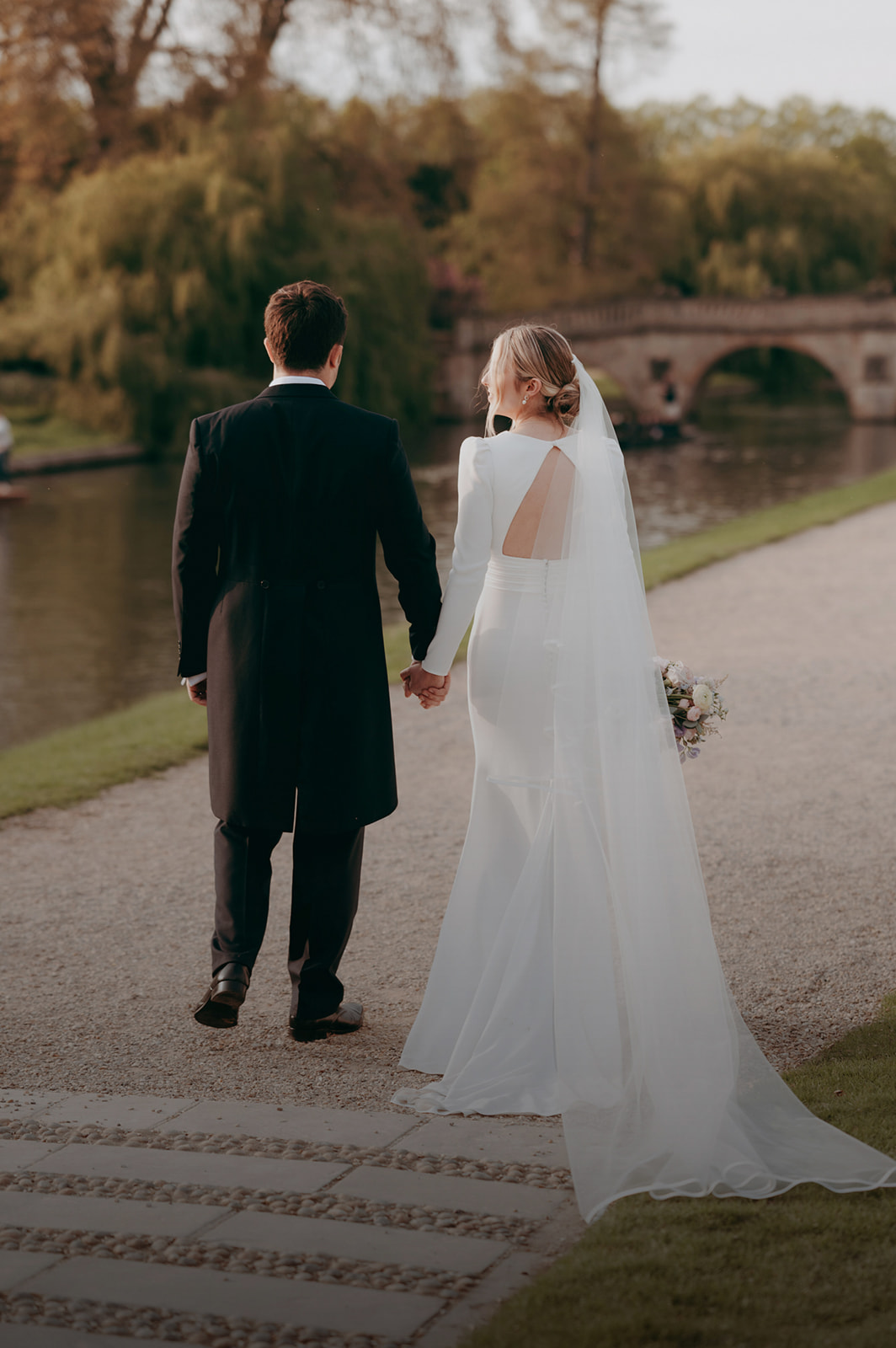 Cambridge groom in a long tailed suit with his bride in a backless dress with a long veil, taken alongside the river Cam by Damien Vickers Photography