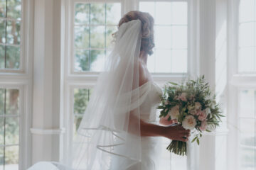 A bride looks towards a bay window as she stands beneath a chandelier holding a bouquet of pink flowers - by Damien Vickers Photography