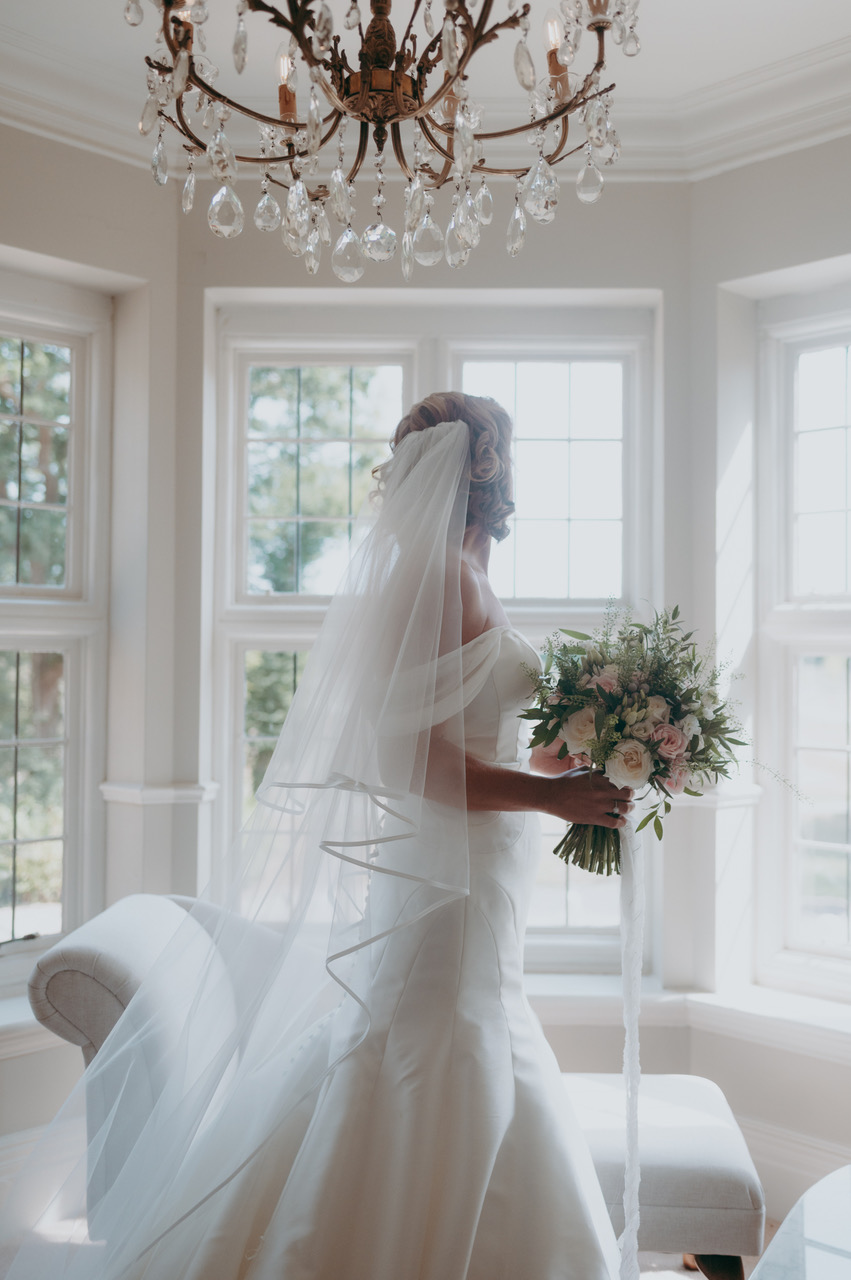 A bride looks towards a bay window as she stands beneath a chandelier holding a bouquet of pink flowers - by Damien Vickers Photography