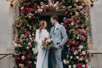 A bride and groom stand before an opulent floral archway with pink and red roses. She's wearing a minimal white dress and he's in a pale blue suit - by Damien Vickers Photography in Cambridge