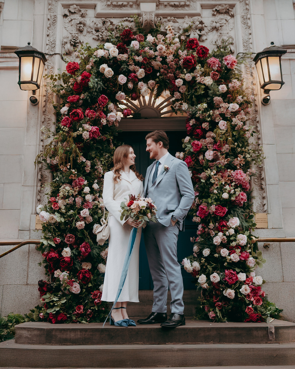 A bride and groom stand before an opulent floral archway with pink and red roses. She's wearing a minimal white dress and he's in a pale blue suit - by Damien Vickers Photography in Cambridge