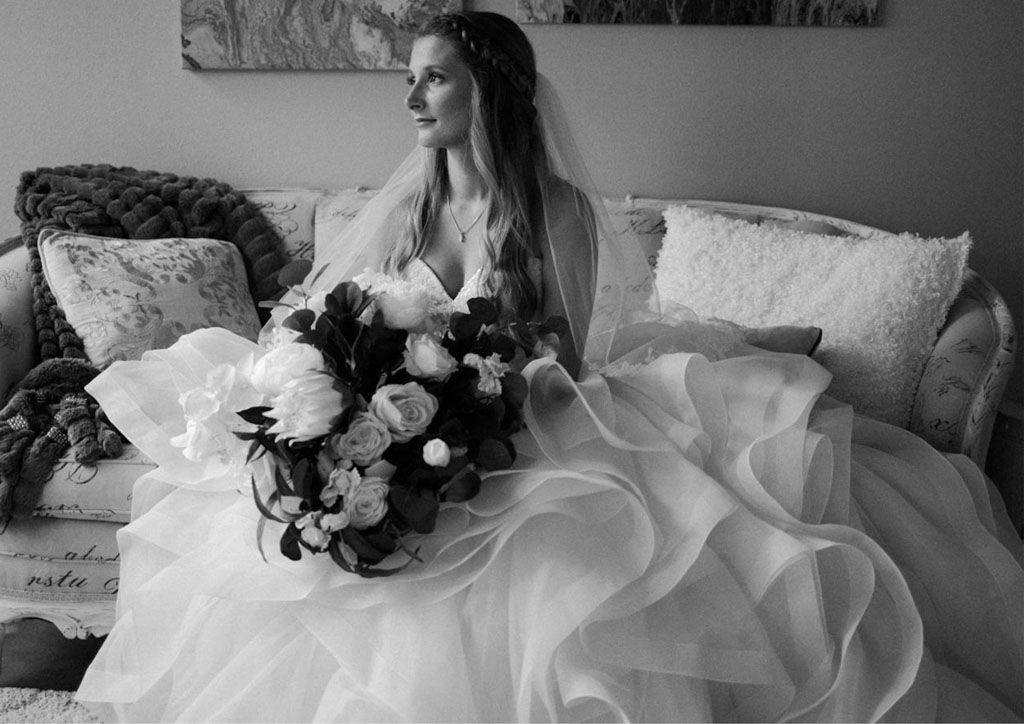Black and white image of a bride sat with her skirts pooled around her, holding a bouquet of flowers