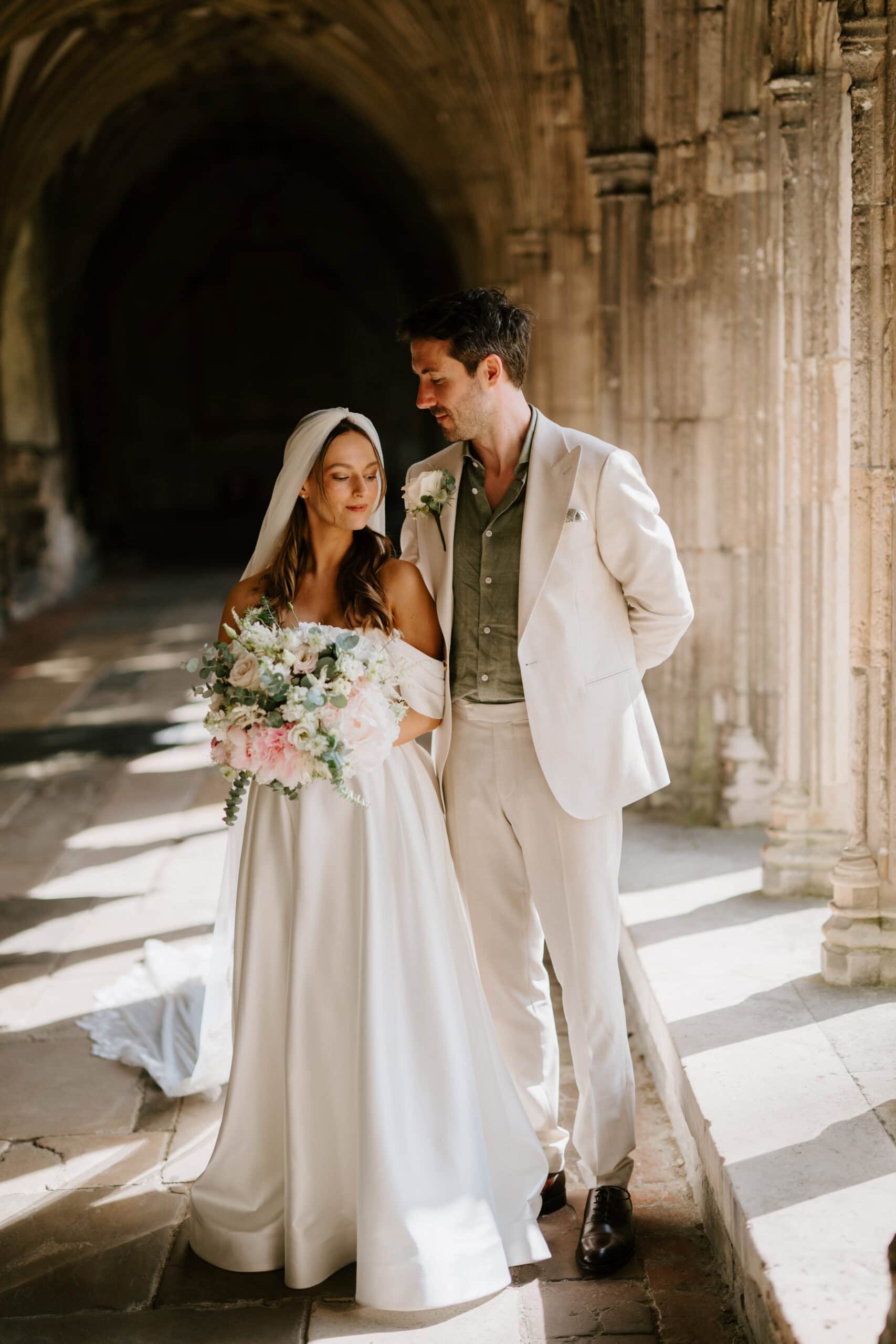Bride and groom stood in sunlight beneath cloistered archways at Canterbury Cathedral by Pearce Wedding Photography in Kent