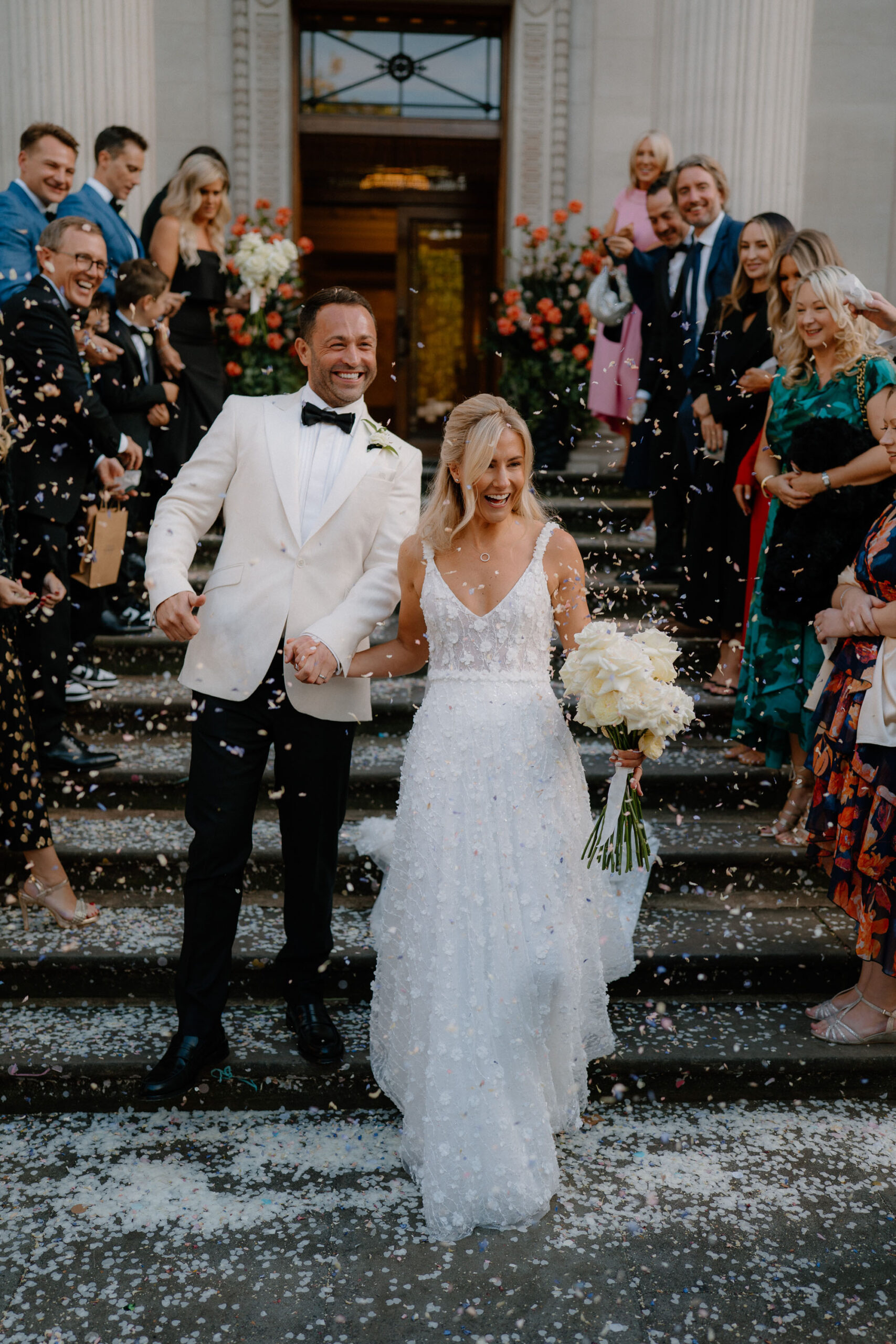 Groom and bride hand in hand at the end of their confetti walk, with white tissue confetti all around and guests smiling - by Tom Aizenberg Photography in London