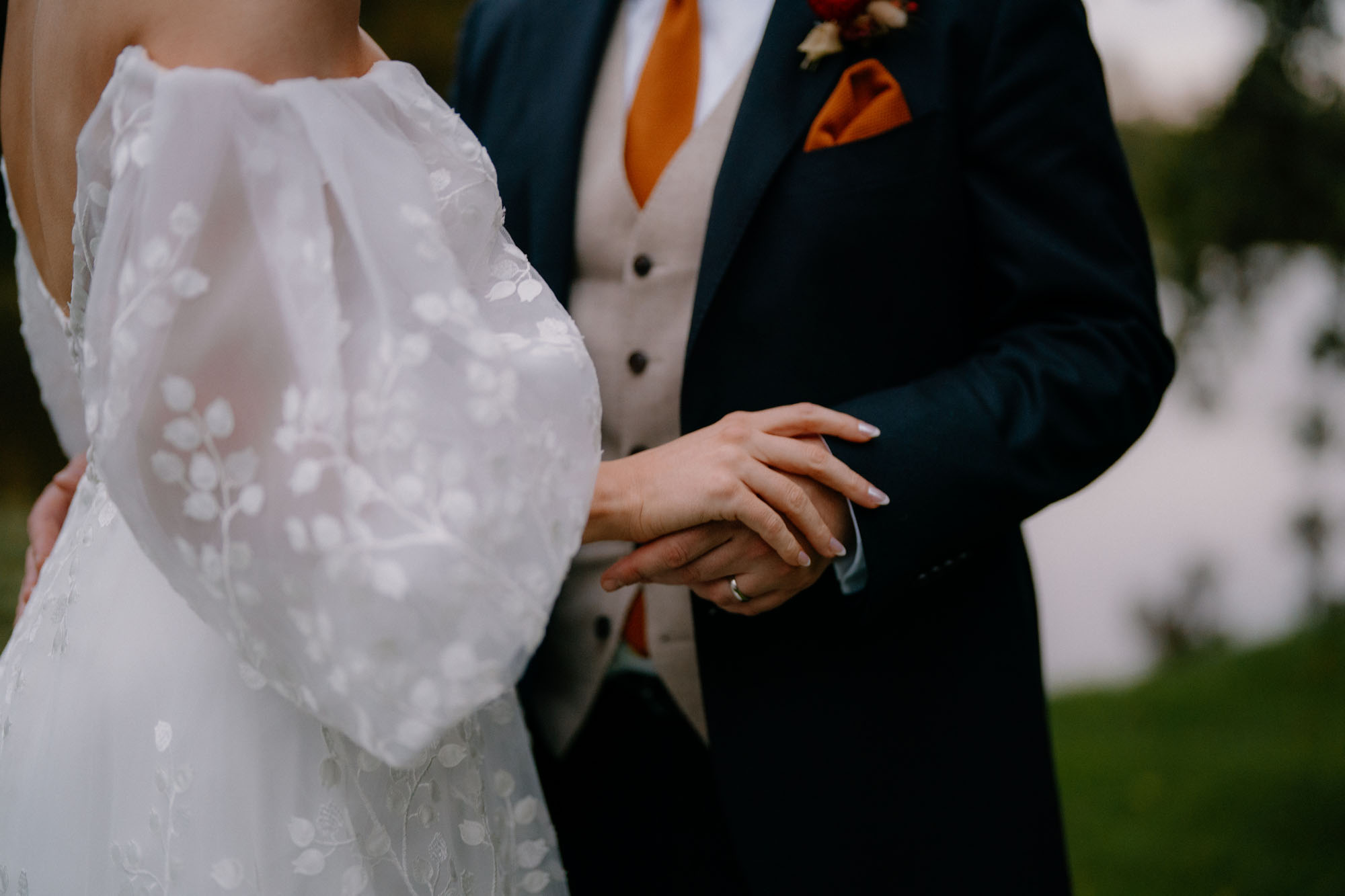 Close up of a newly married couple holding hands. The bride is wearing a white dress with embroidered sleeves and the groom's in a suit with waistcoat and orange tie - by Tom Aizenberg in London