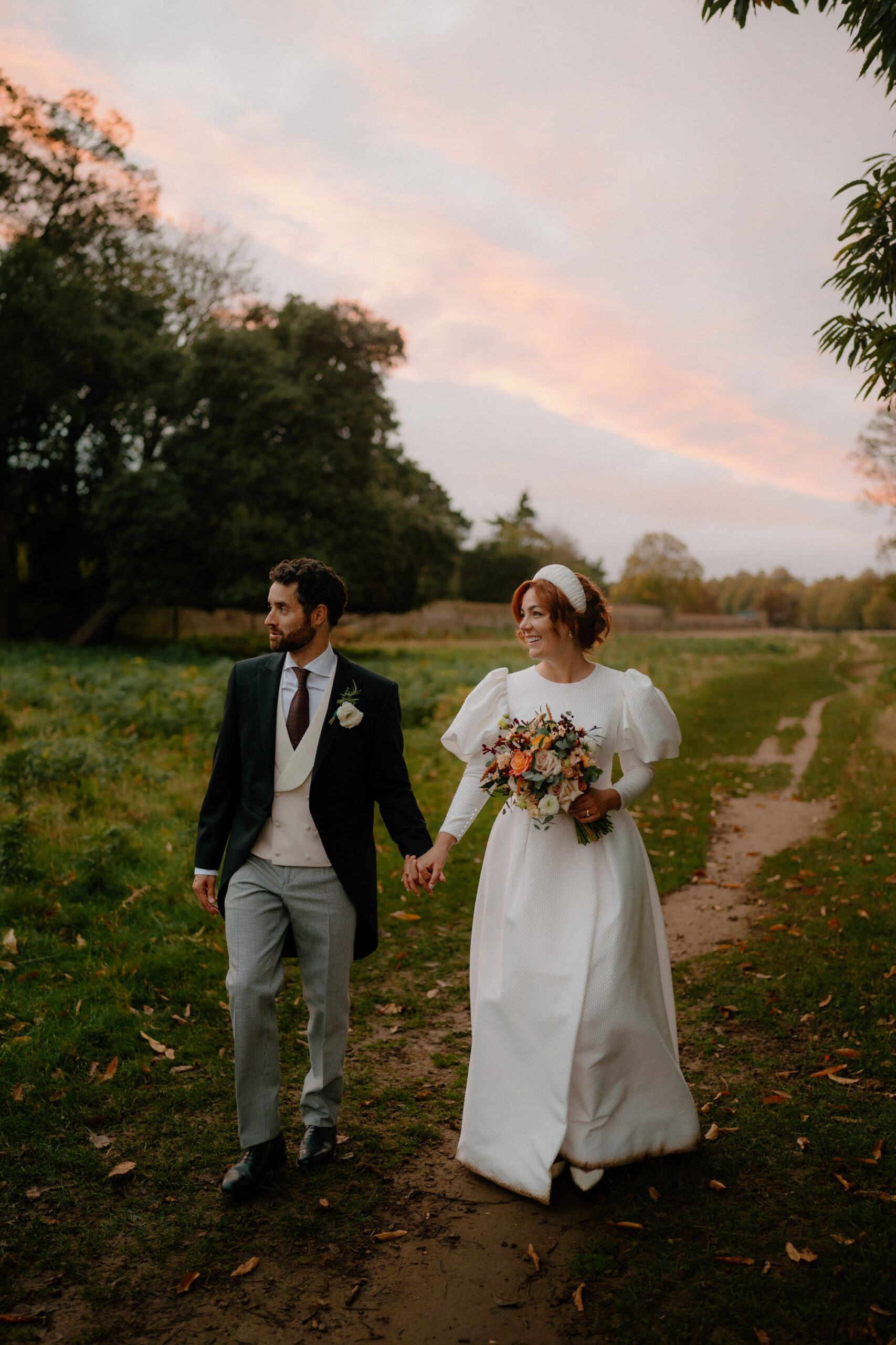 A groom and bride walking hand in hand with a delicate sunset in the sky behind them. Documentary wedding photography by Tom Aizenberg