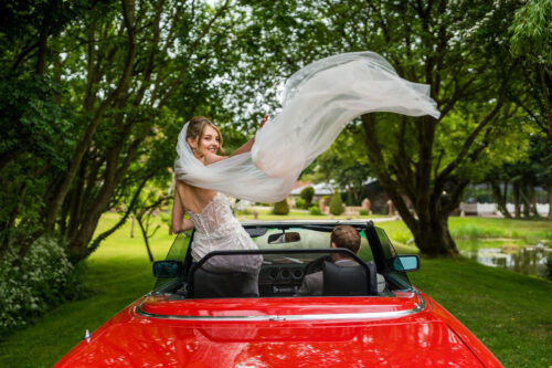 A bride stands in a sports car with the roof open, her veil catching the wind at Solton Manor - Benjamin Toms Photography | Kent