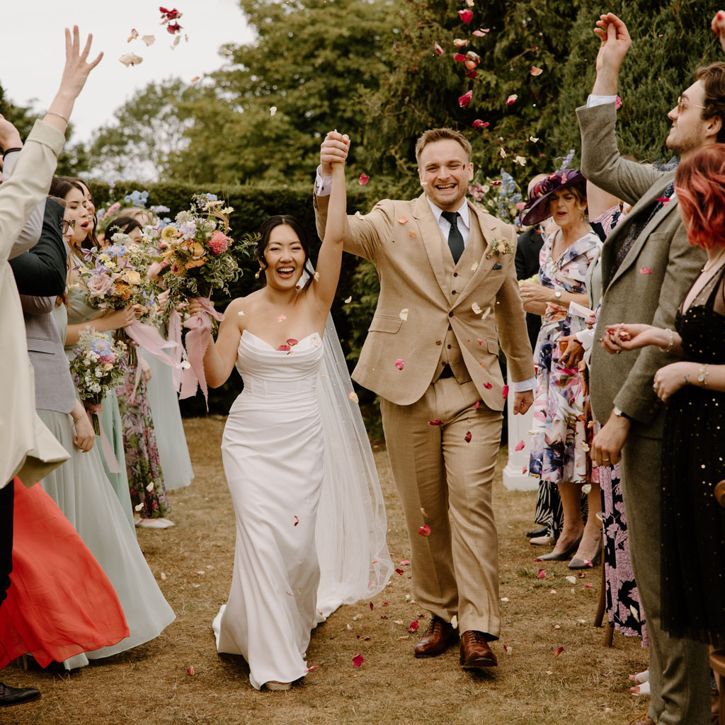 newlyweds walk with hands held and raised through a shower of biodegradable confetti from The Dried Petal Company | image credit Soul Capture Photography