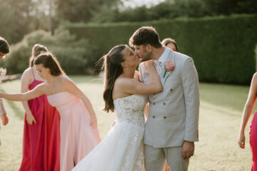 Groom and bride steal a kiss as they stand on a sunny lawn with bridesmaids in pink behind them | Pearce Wedding Photography at The Old Kent Barn