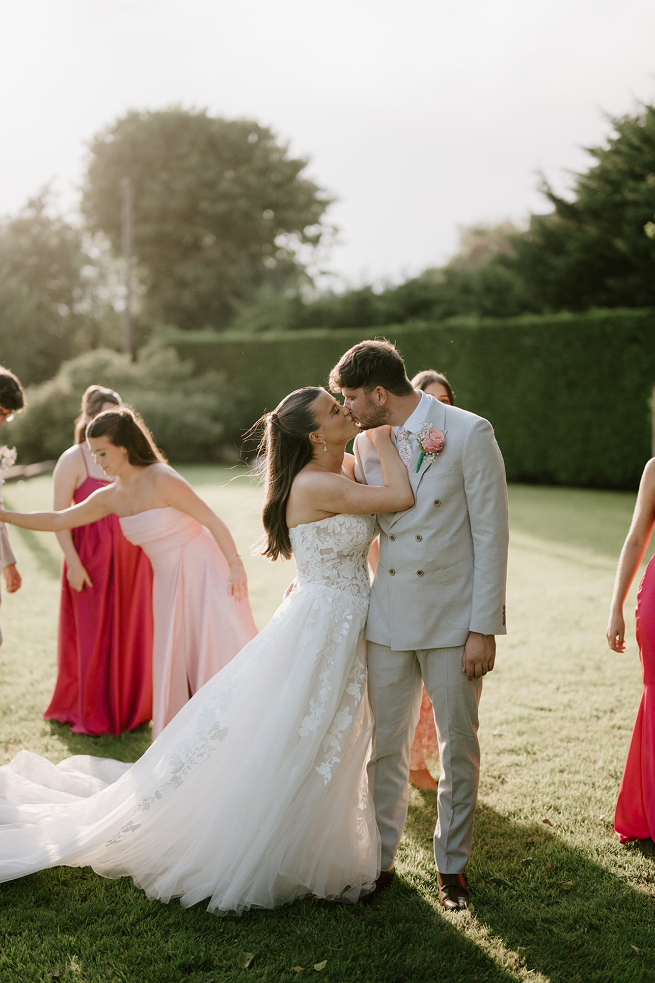 Groom and bride steal a kiss as they stand on a sunny lawn with bridesmaids in pink behind them | Pearce Wedding Photography at The Old Kent Barn