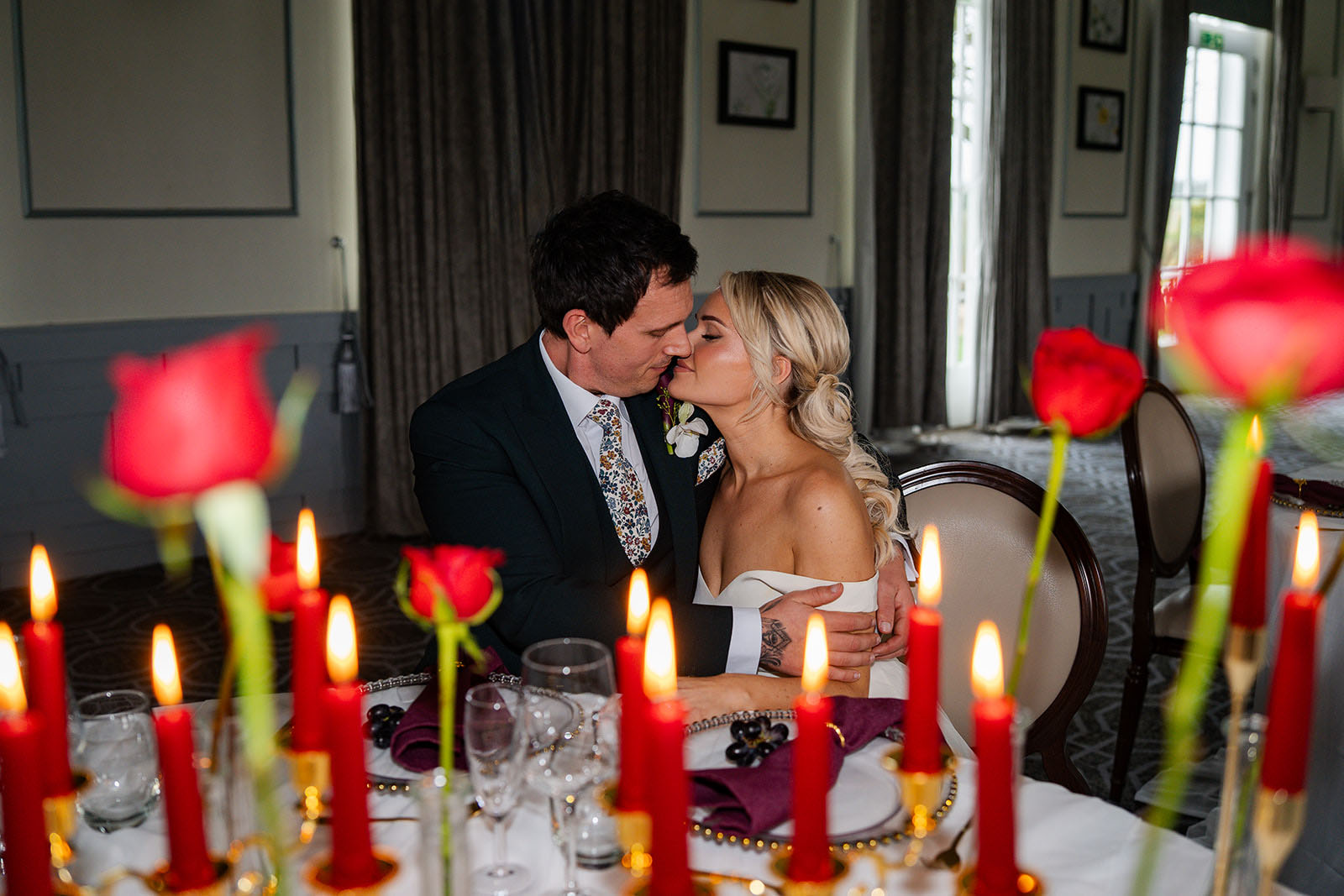 A couple kiss in the background | in the foreground, a wedding table styled with lit red candles by Lauren Batten Events