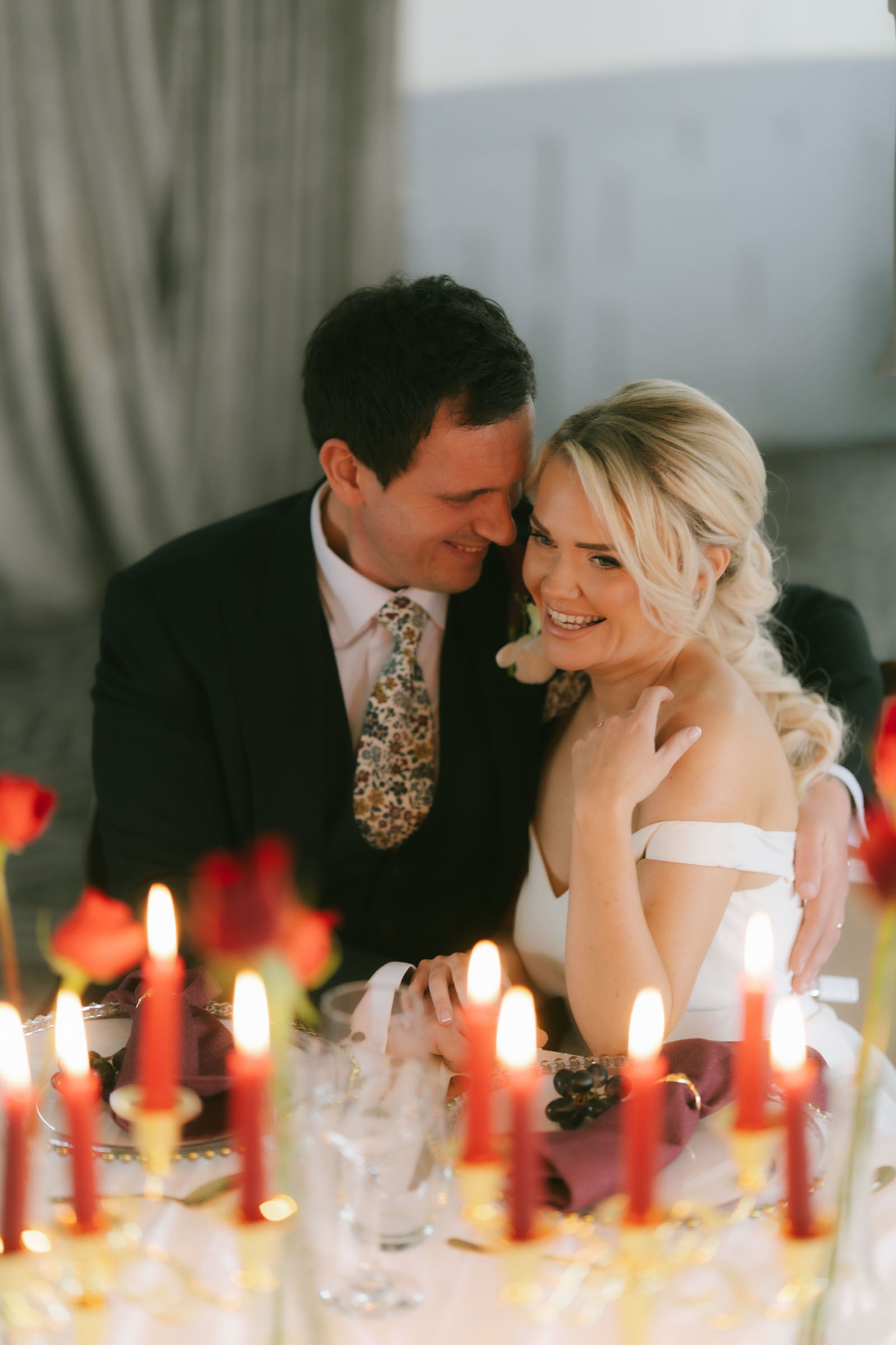 A man in a wedding suit has his arm around a smiling woman in a wedding dress. There are lit red candles in the foreground by Lauren Batten Events