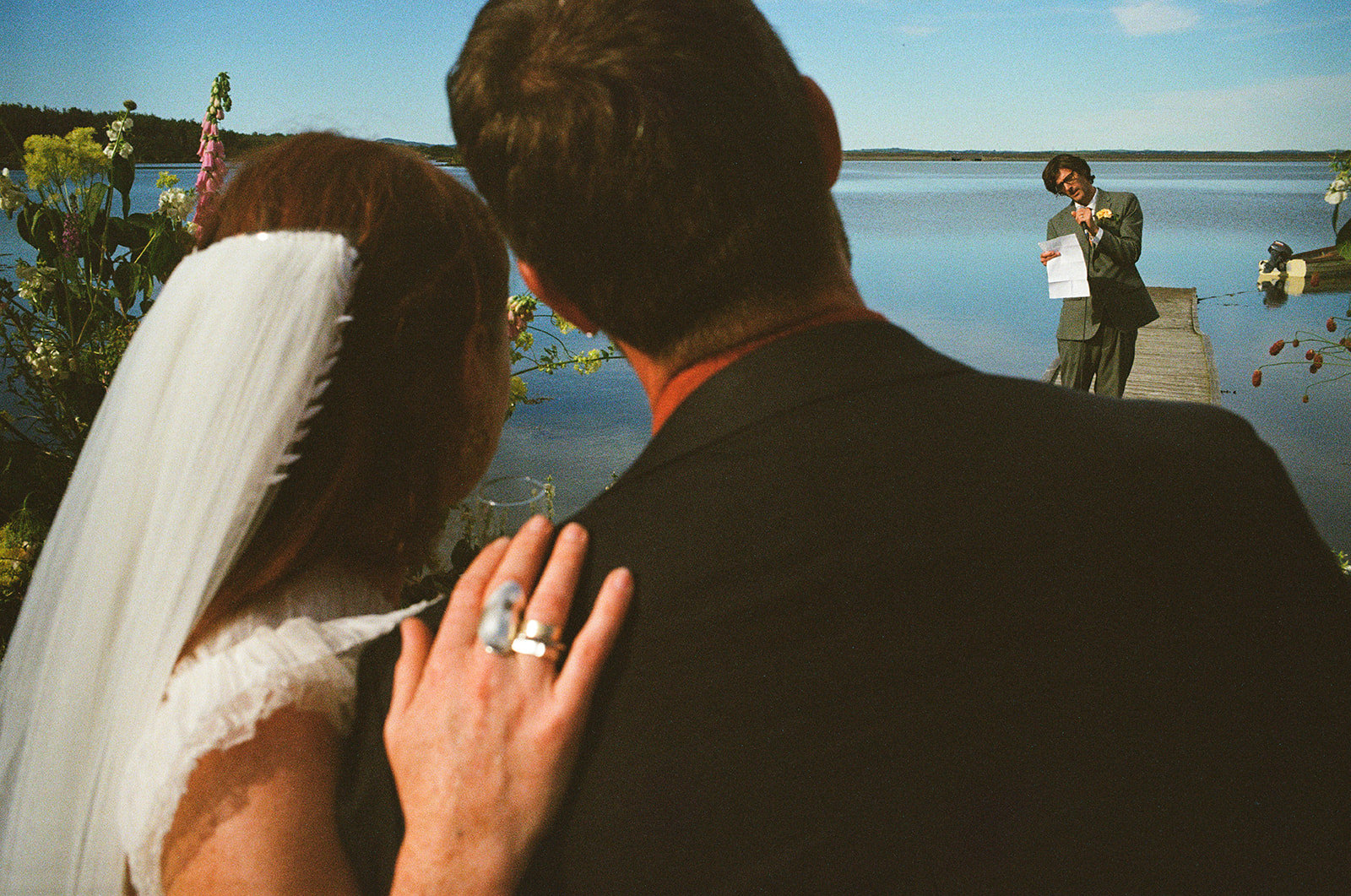 Newlyweds looking seaward as a man reads a wedding speech. The bride's hand is on the groom's shoulder. Analogue film wedding photography by York Place Studios