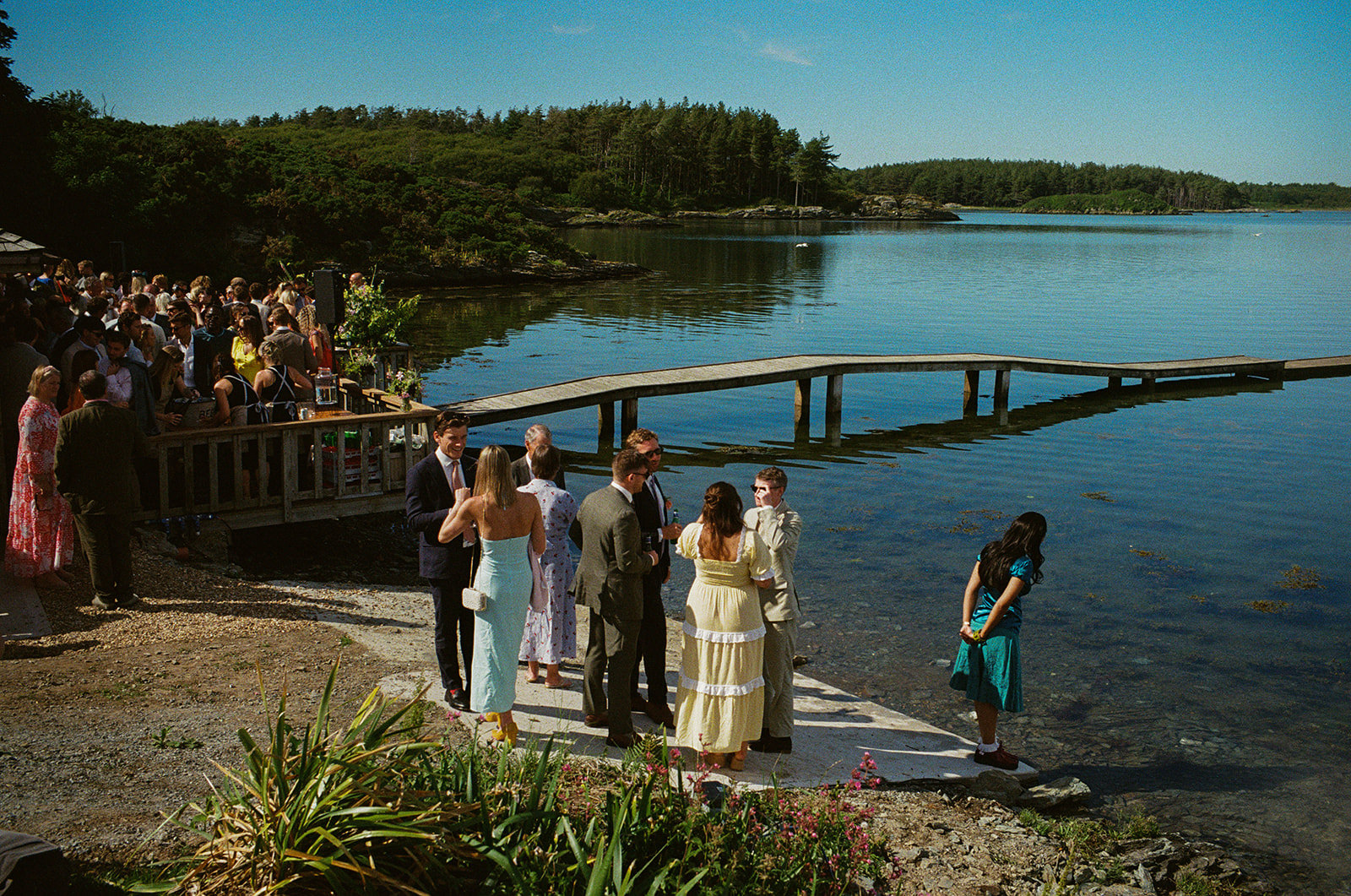 Lakeside wedding photography with a jetty in the background. Guests gather on a small beach. Film wedding photography by York Place Studios