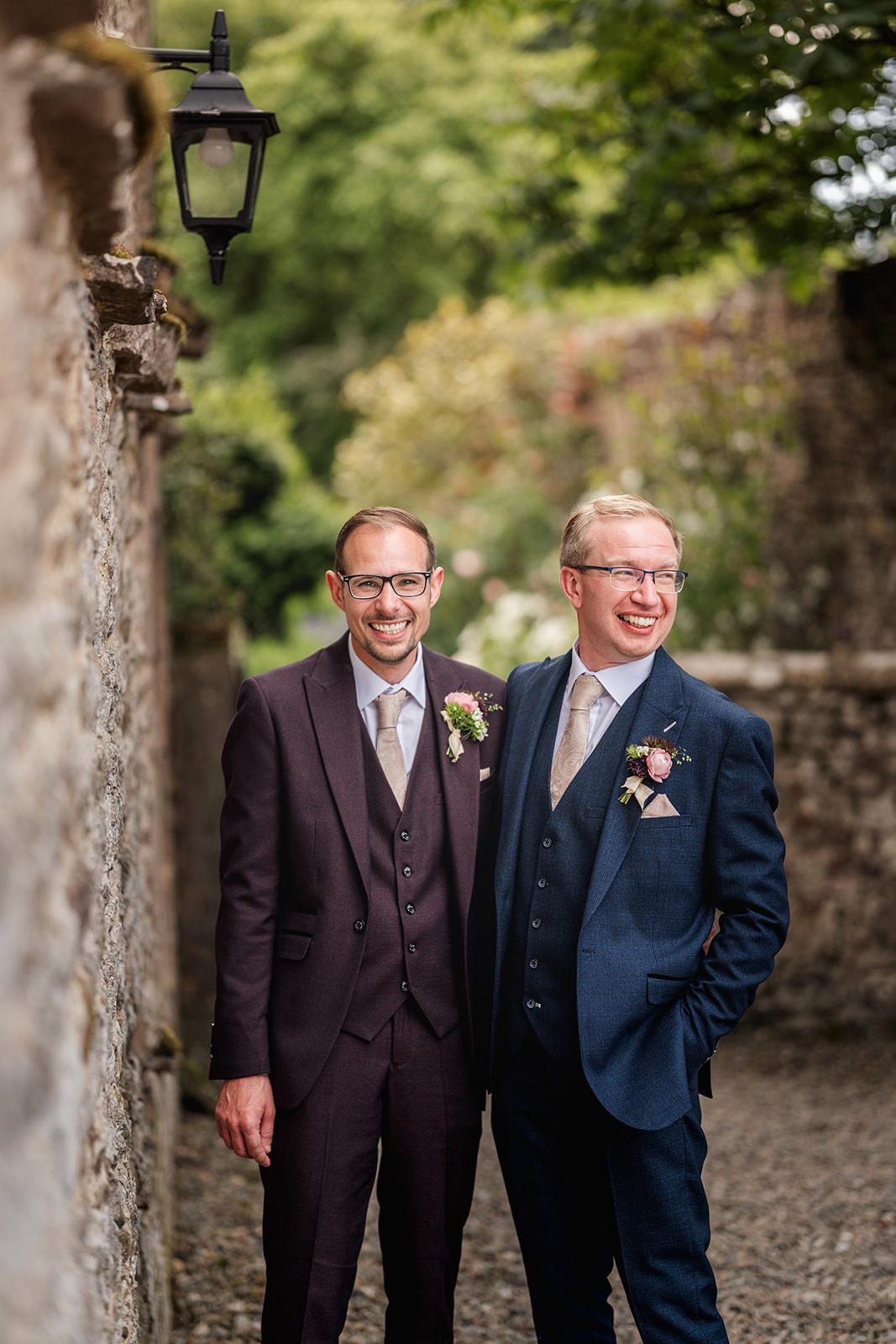 Two grooms beside a barn wall with a lantern, with a garden in the background | Jaye Peg Photography at Holesfoot Cumbria