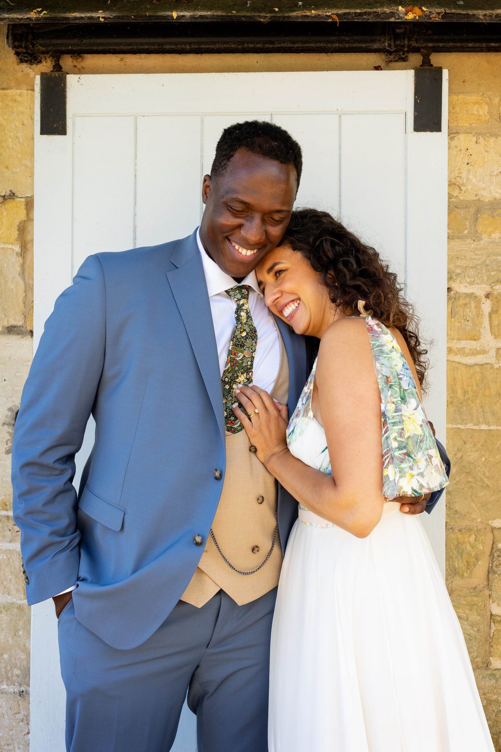 Groom and bride cuddling and smiling by a white barn door. He's in a blue suit and waistcoat, she's in a painted wedding dress | Martin Dabek Photography at Ashley Wood Farm Wiltshire