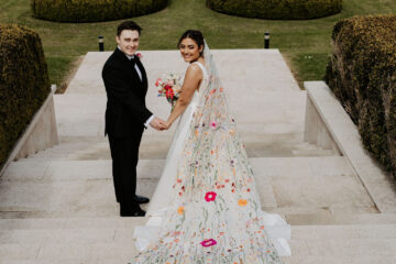 groom and bride on steps leading towards landscaped gardens. She's wearing a very long veil embroidered with colourful flowers | Hannah Timm Photography