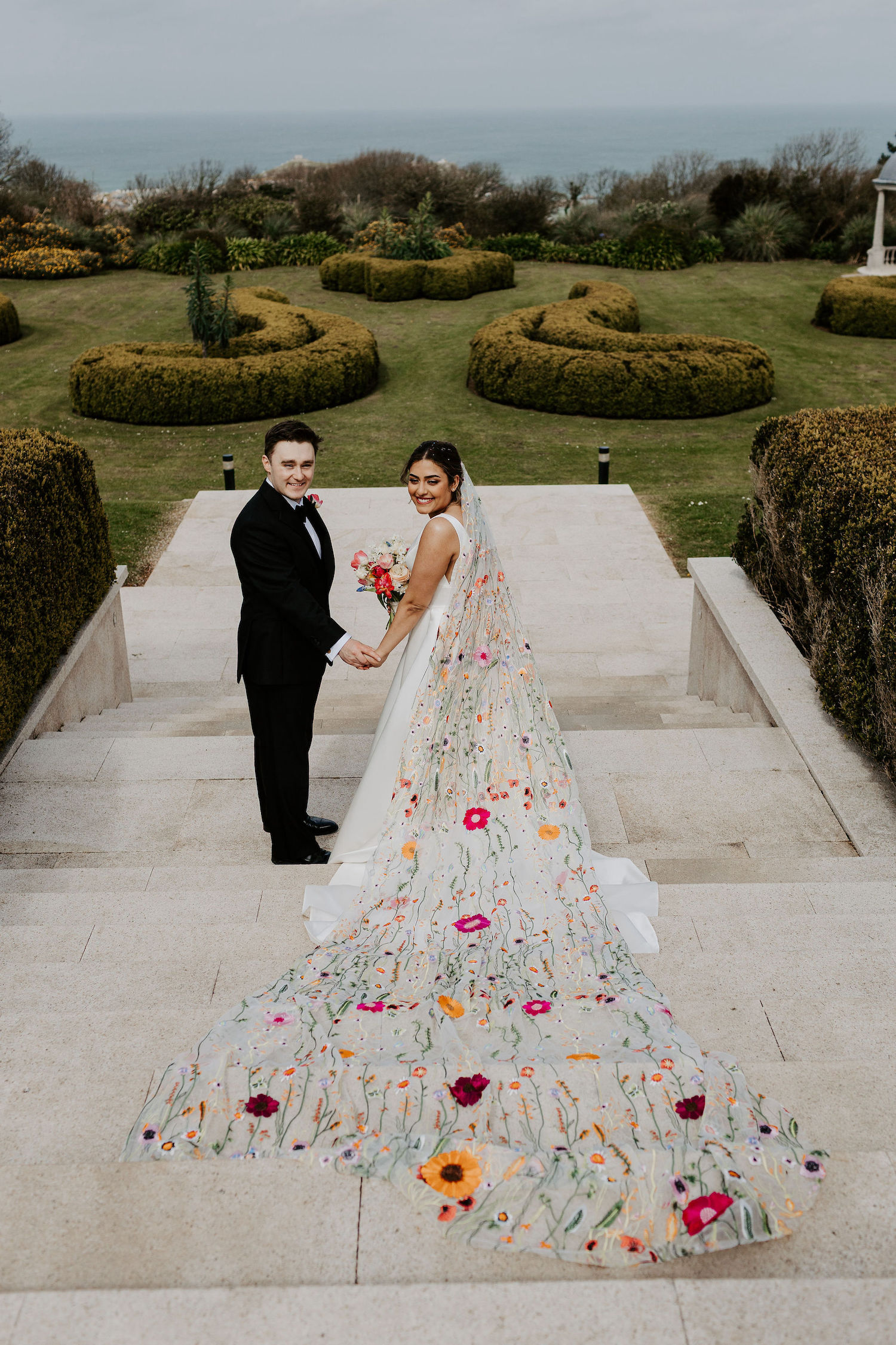 groom and bride on steps leading towards landscaped gardens. She's wearing a very long veil embroidered with colourful flowers | Hannah Timm Photography