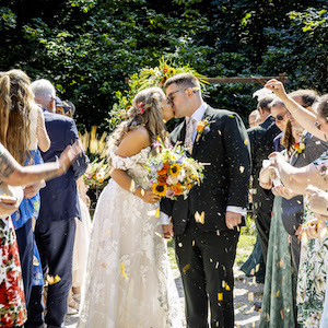 Bride and groom kissing while guests throw flower confetti by Ezra's Confetti